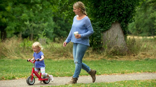 Woman walking along a path with a small child on a scooter, in the grounds of Morden Hall Park, London.